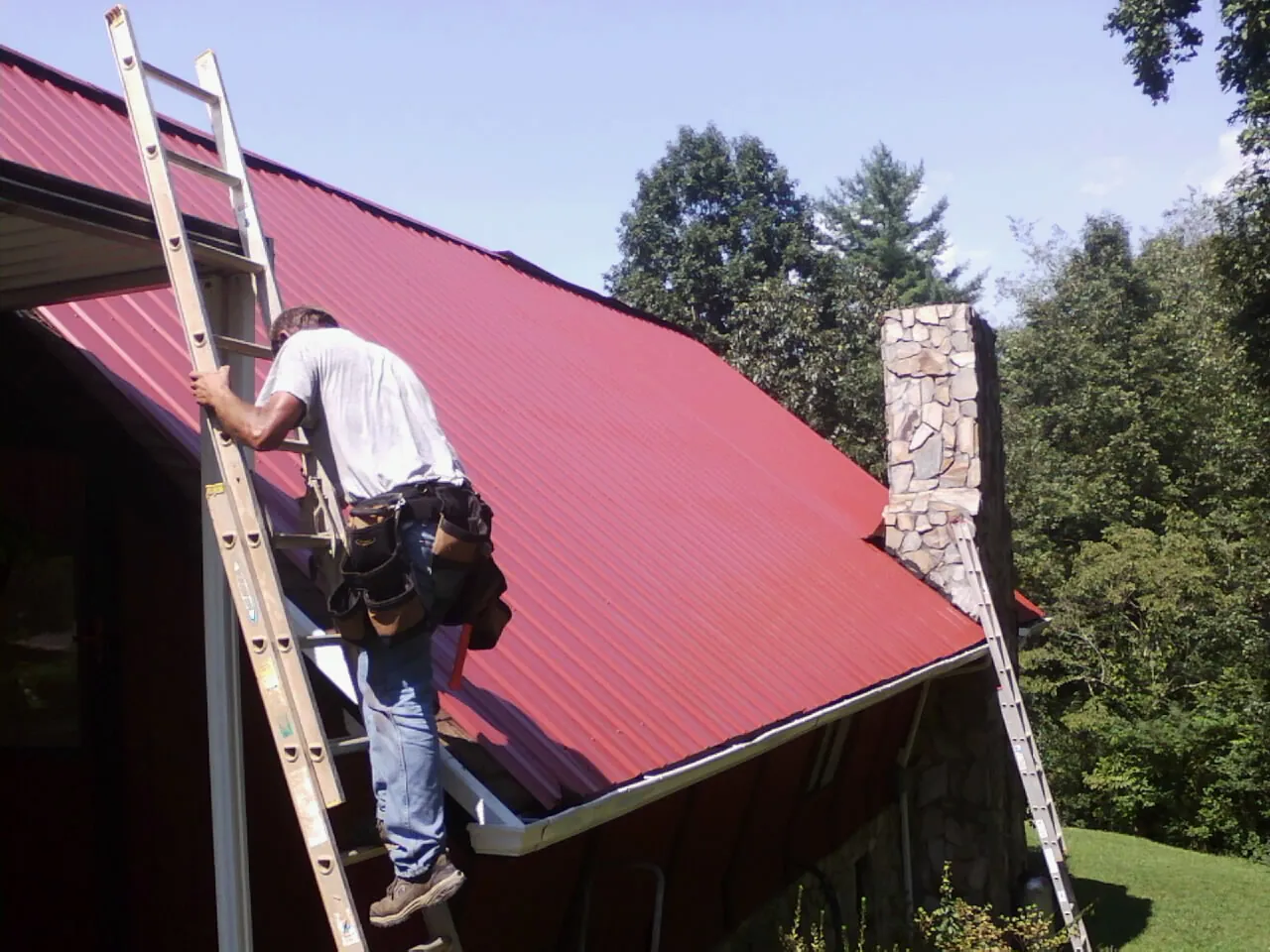 Roofer on a ladder beside a red metal roof and stone chimney — Soffit & Fascia Repair by Roofing Brothers in Shiner